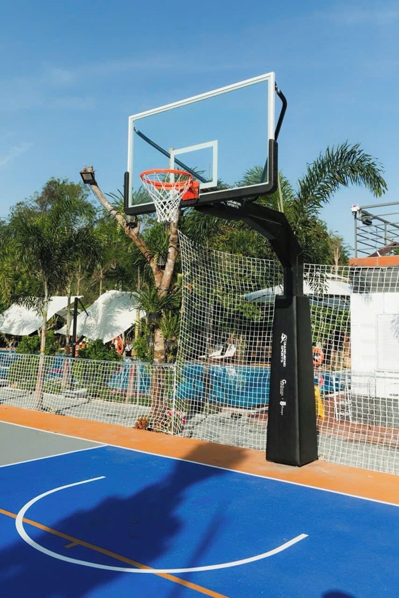 An outdoor basketball court with a netted enclosure, surrounded by palm trees and a blue playing surface.