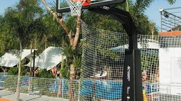 An outdoor basketball court with a netted enclosure, surrounded by palm trees and a blue playing surface.