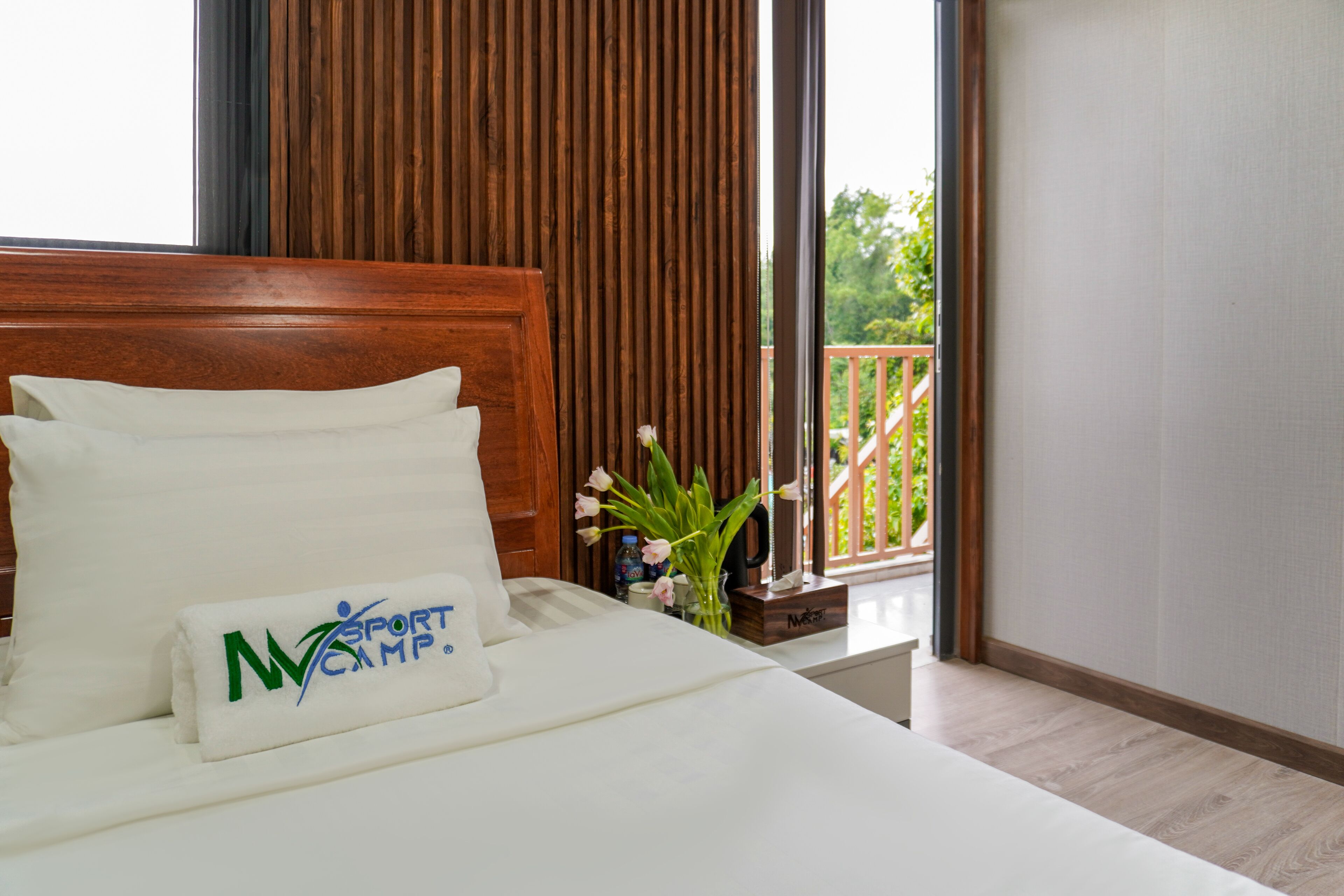 A hotel room with a wooden headboard, a bed with white linens, a small table with a vase of flowers, and a balcony with a wooden railing.