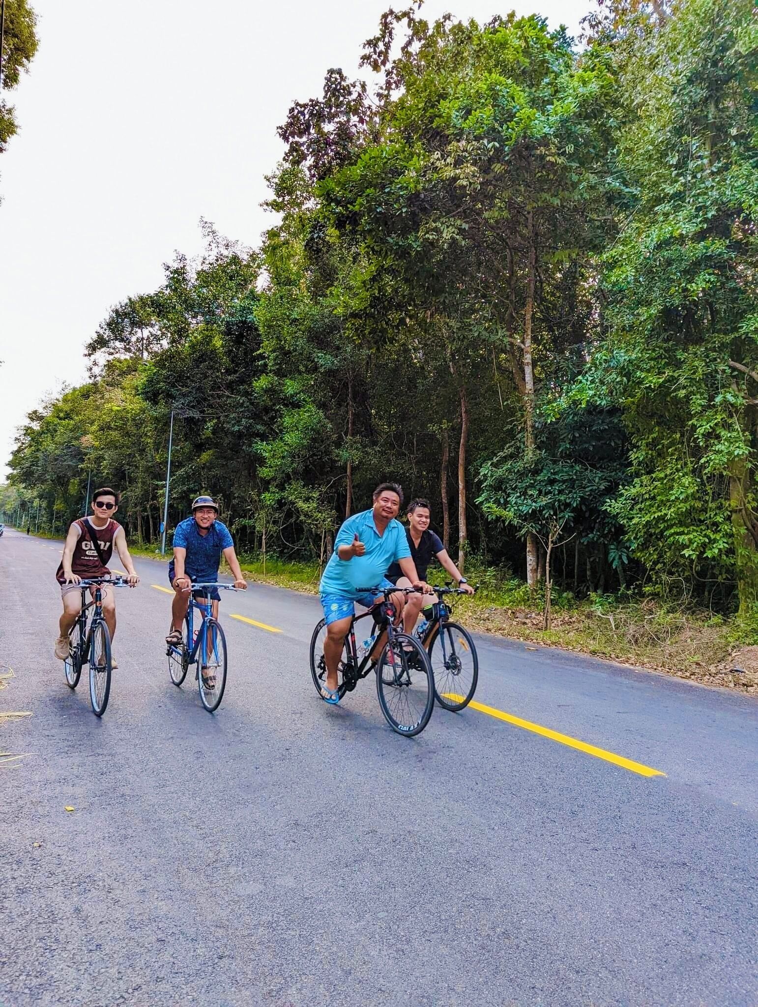 Four people riding bicycles on a paved road surrounded by dense greenery.