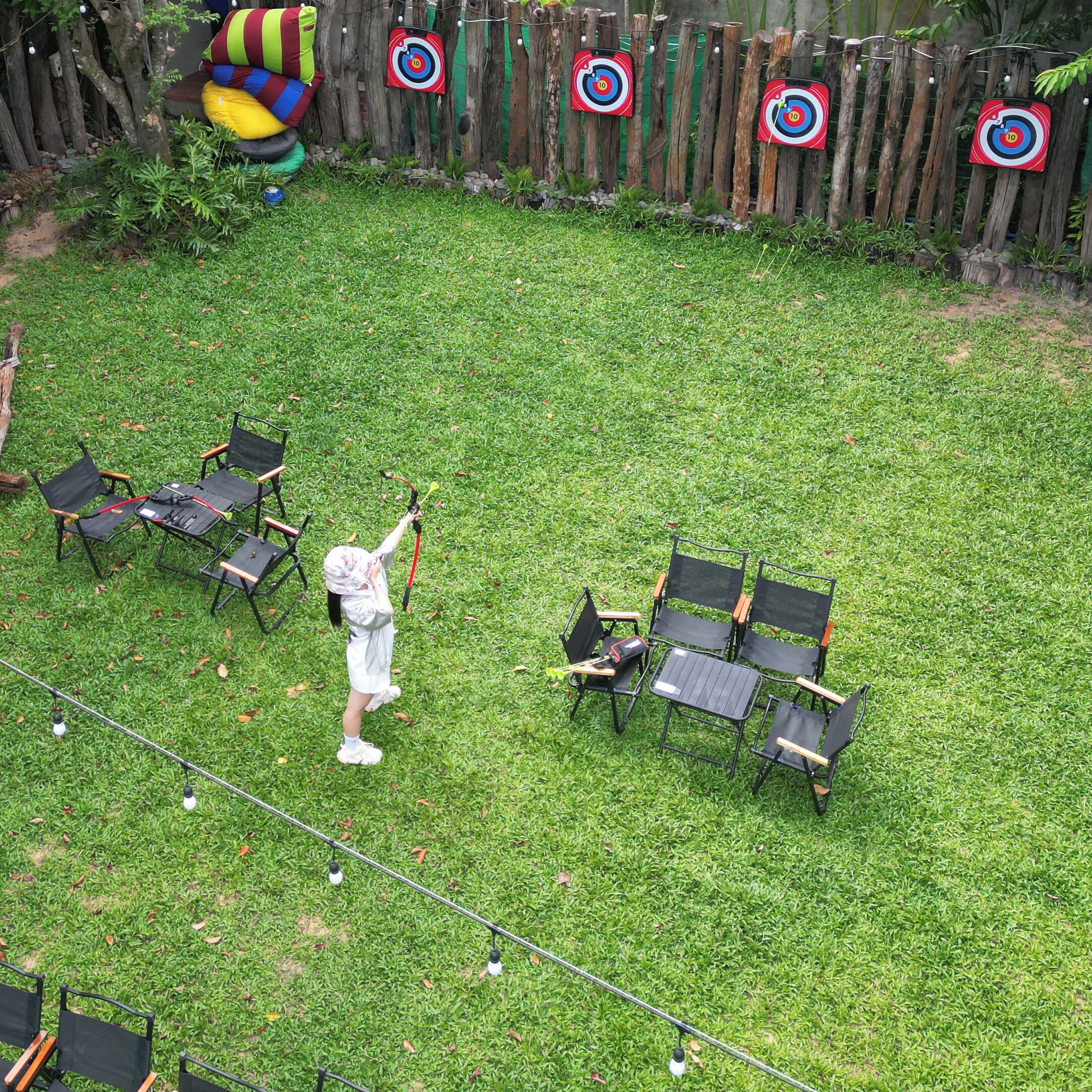 A grassy outdoor area with a target practice setup, including a child aiming a slingshot, chairs, and a wooden fence.