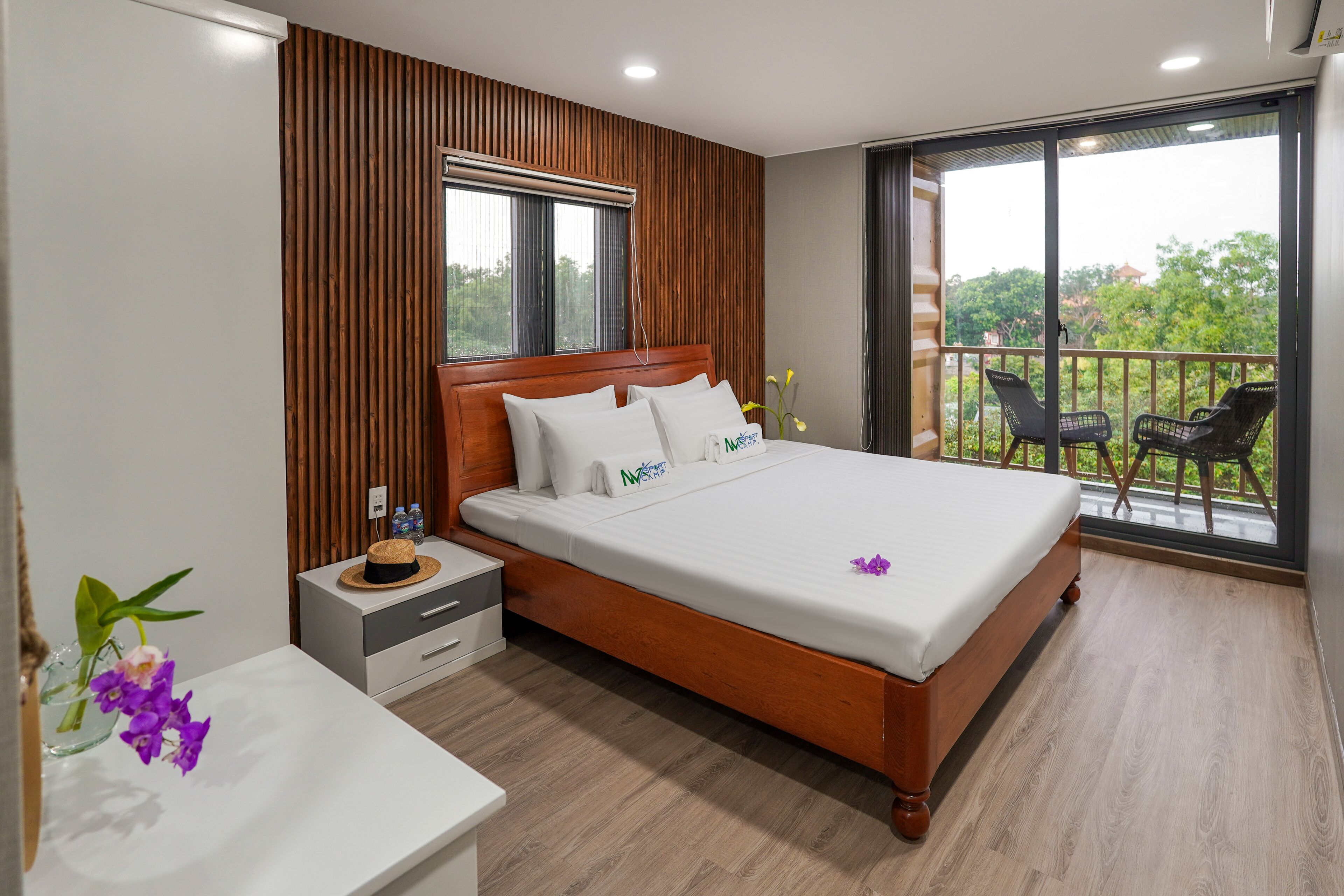 A modern bedroom with a wooden headboard, a large bed, a nightstand, a small table with a vase of flowers, and a balcony with chairs.