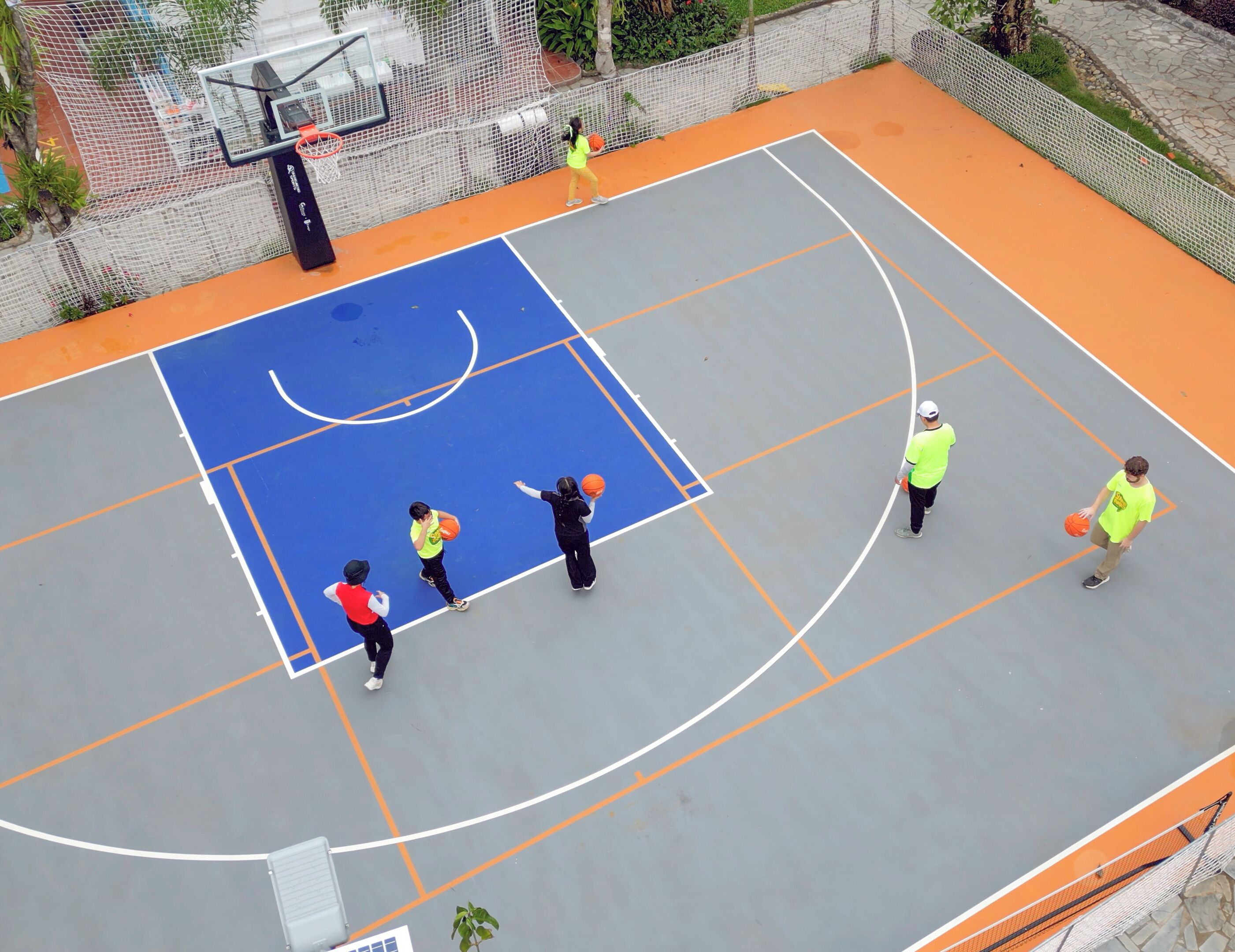 An outdoor basketball court with a blue and orange playing surface, surrounded by a white fence.