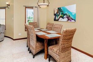 Een eetgedeelte met een houten tafel en rieten stoelen, een schilderij aan de muur en een raam met gordijnen.