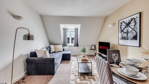 A modern living room with a sofa, coffee table, and dining area. A TV is mounted on the wall, and a window provides natural light.
