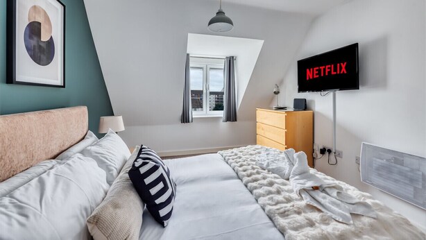 A modern bedroom with a bed, a TV displaying 'NETFLIX', a window, and a ceiling light.
