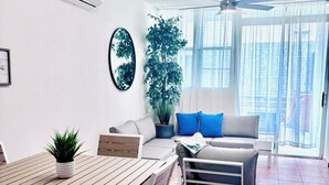 A modern living room with a wooden dining table, white furniture, a large potted plant, a ceiling fan, and a view through large windows. - Beachside With Private Jacuzzy, Full A/c, Totally Renovated, Brand new (Luquillo)