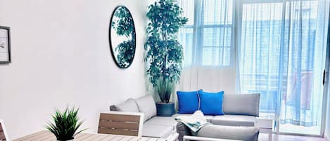 A modern living room with a wooden dining table, white furniture, a large potted plant, a ceiling fan, and a view through large windows.