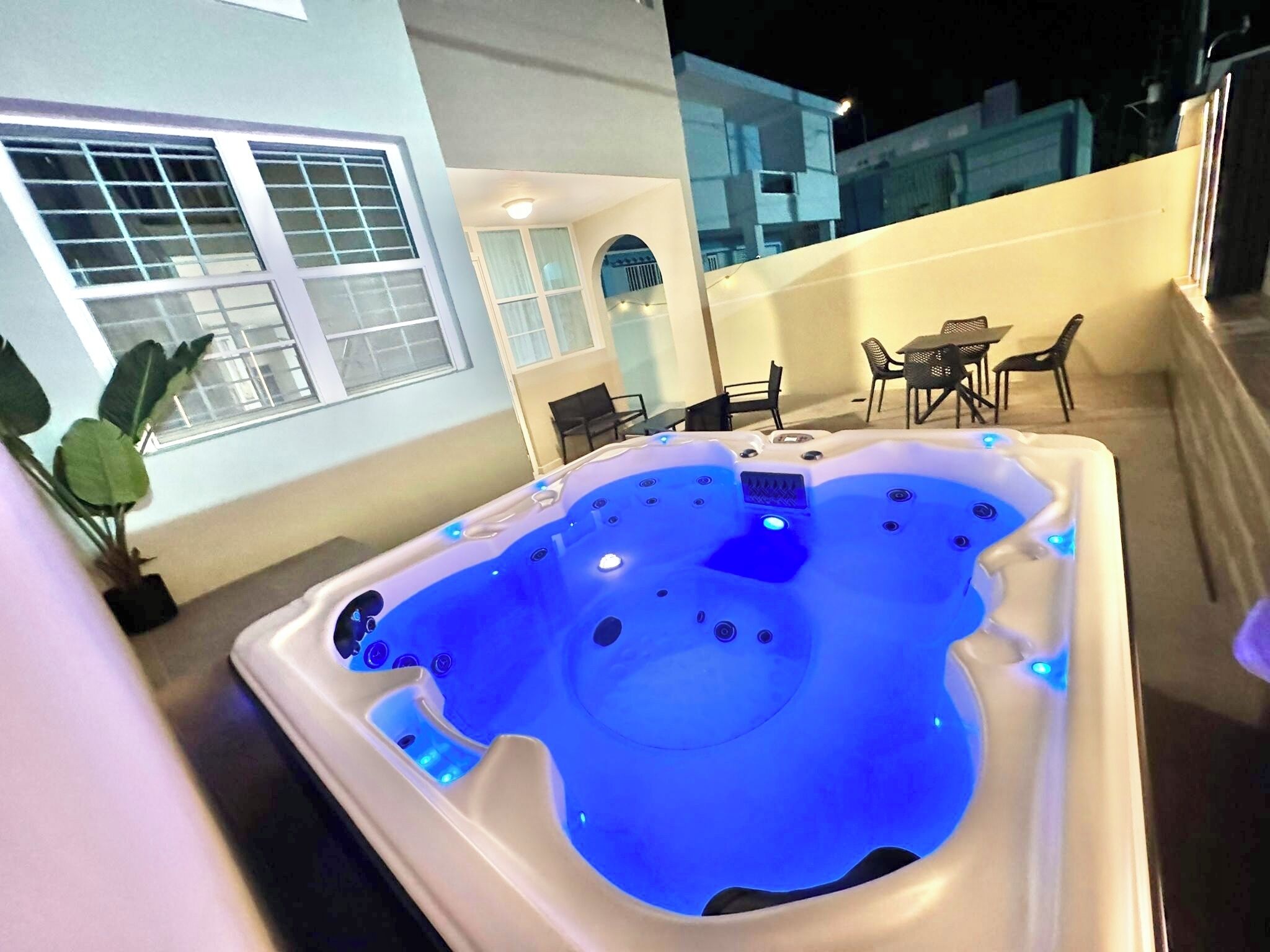 A hot tub with blue lighting, a potted plant, and a patio area with chairs and a table.