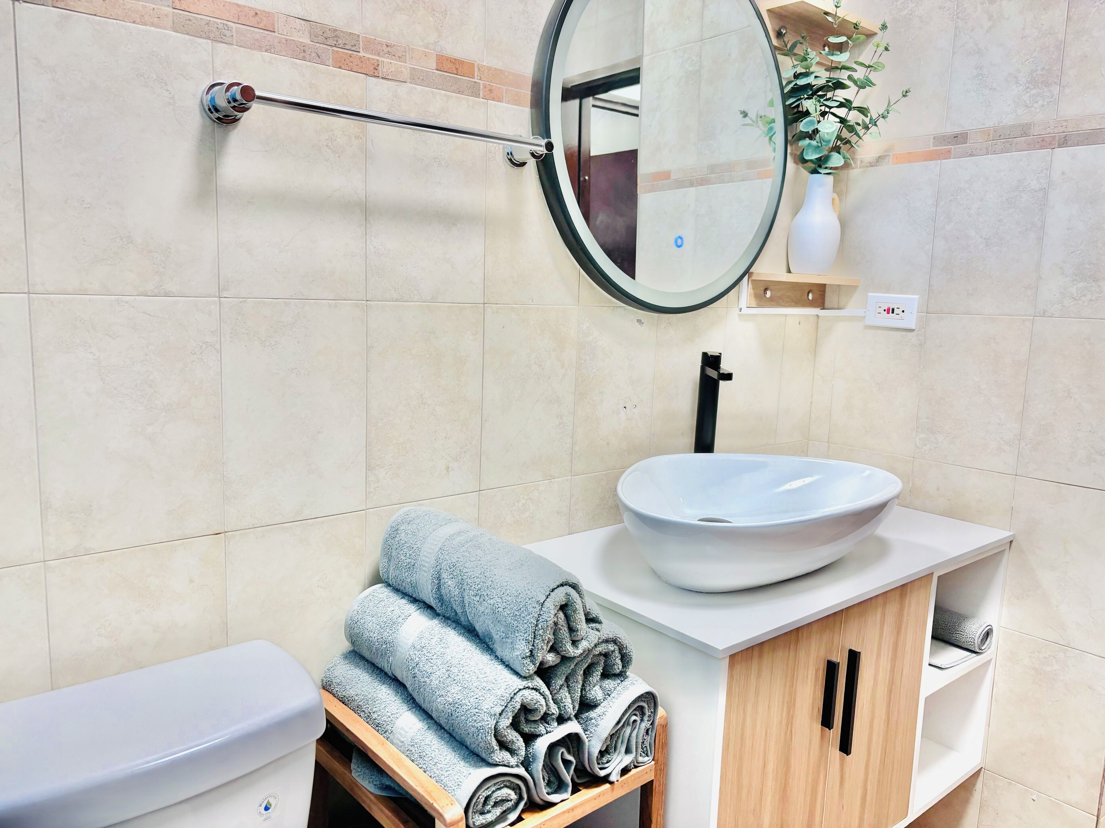 Ein Badezimmer mit einem weißen Waschbecken, einer Toilette, einem Handtuchhalter mit gefalteten Handtüchern, einem Spiegel und einer kleinen Pflanze an der Wand.