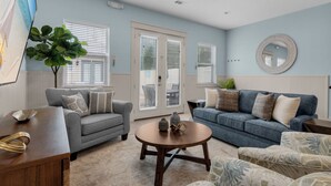 A living room with two sofas, a coffee table, and a TV. There are windows with blinds, a plant, and a mirror on the wall.