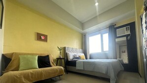 A room with a sofa, a bed, a small table with a plant, and a window with curtains.