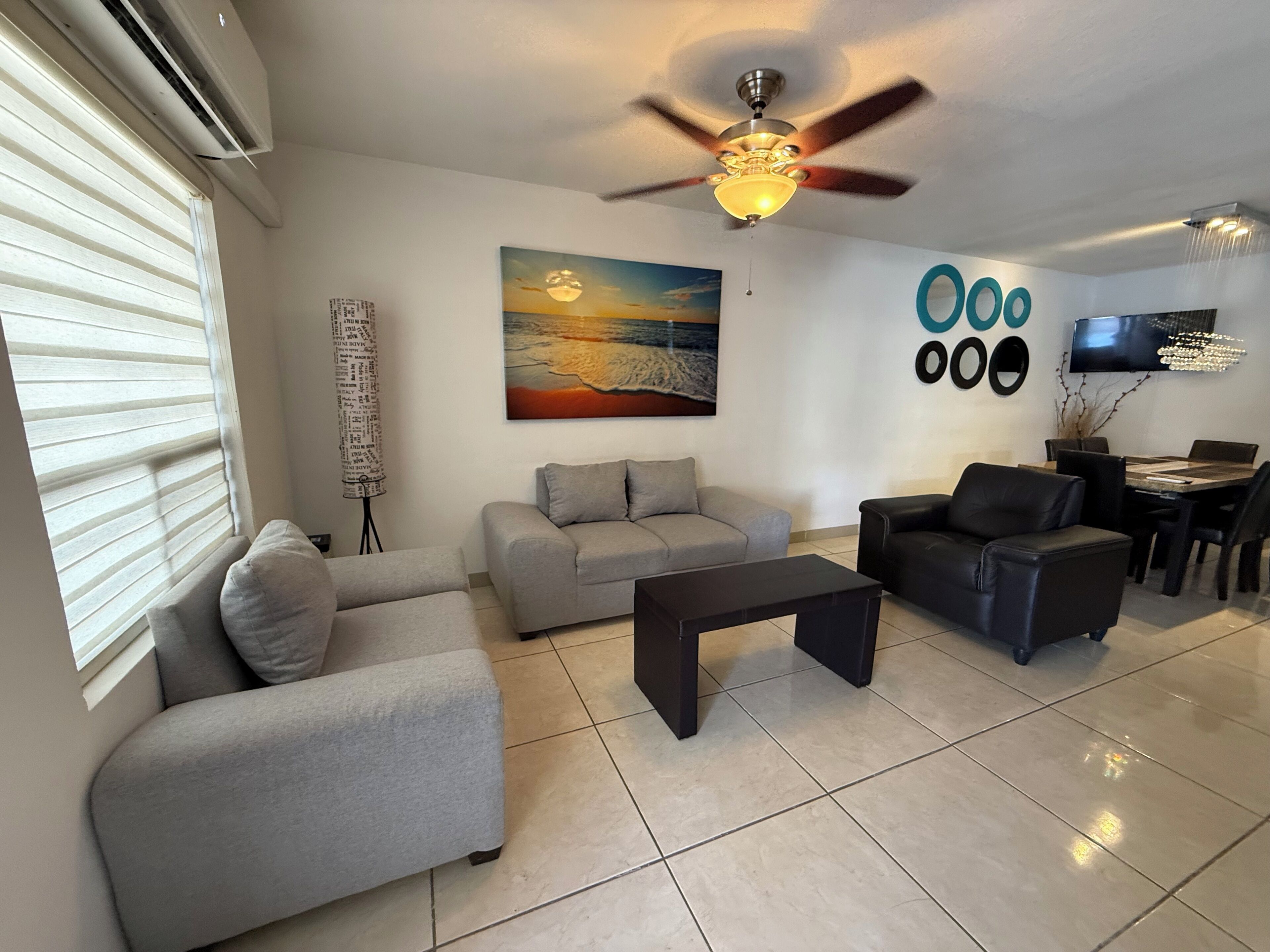 A modern living room with a ceiling fan, a painting, a dining area with chairs, and a TV mounted on the wall.