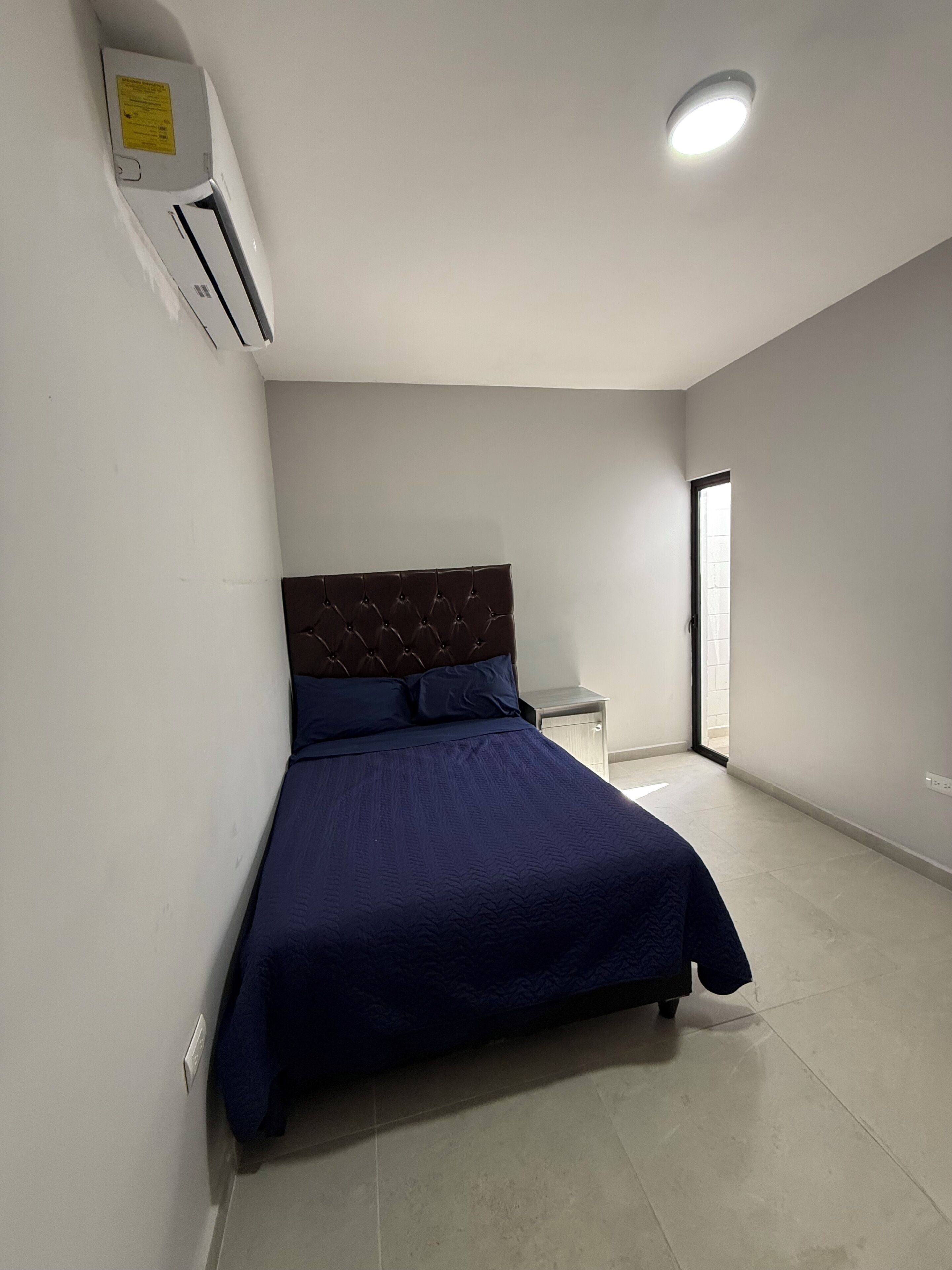 A bedroom with a bed, a nightstand, an air conditioning unit, and a ceiling light.