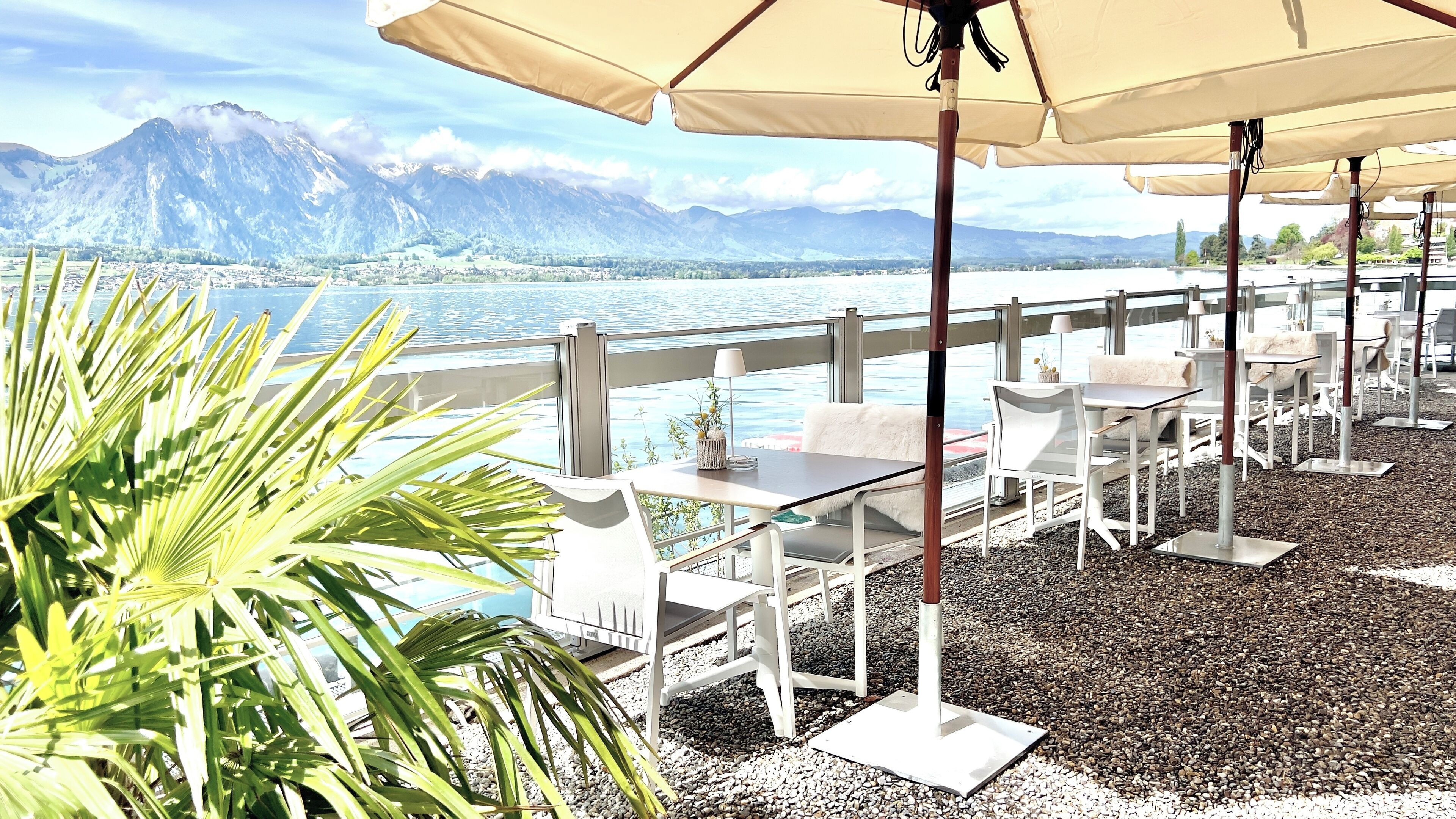 An outdoor terrace with white tables and chairs, beige umbrellas, and a view of a lake with mountains in the background.