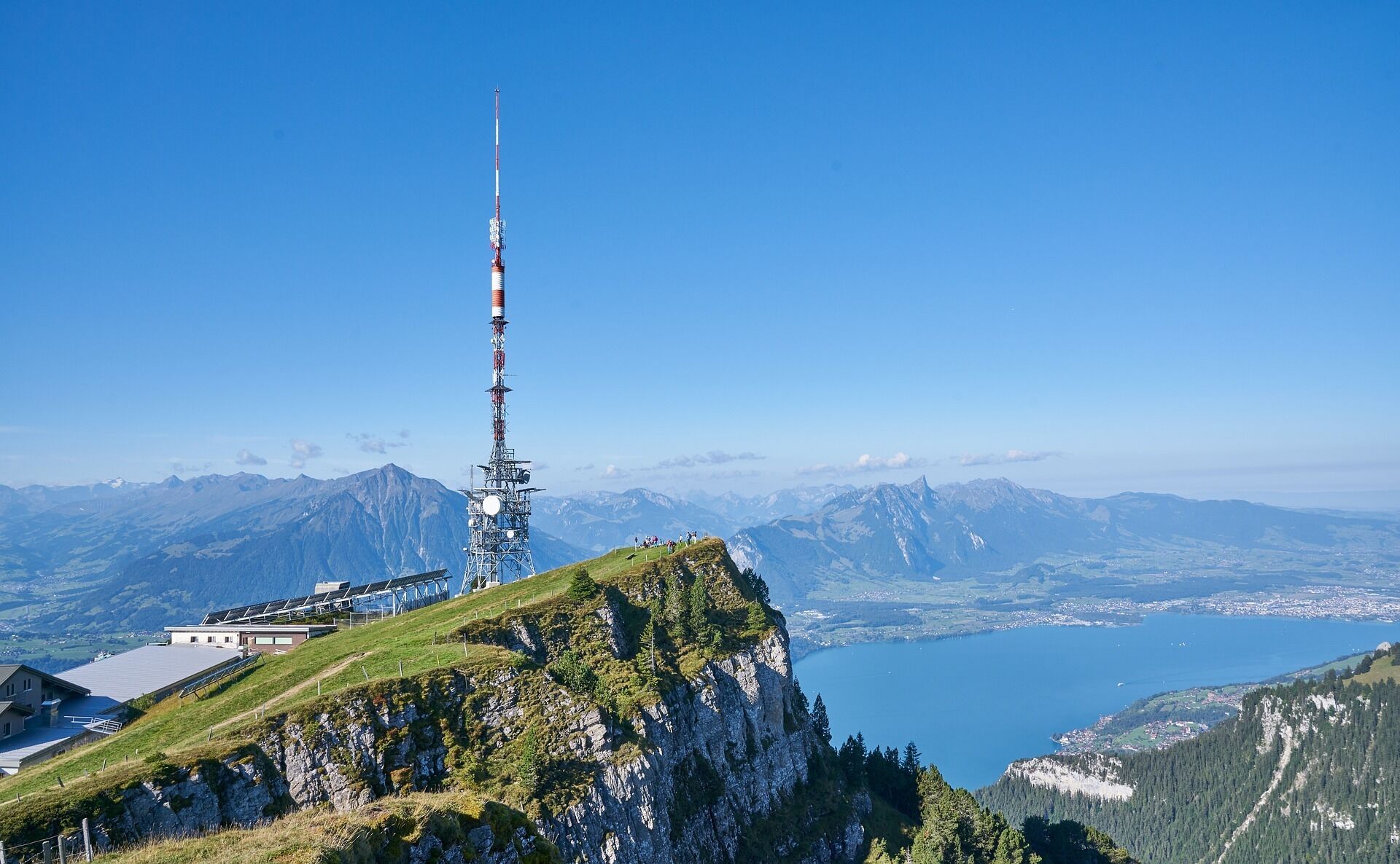 A mountain peak with a tall communication tower, a building with a sloped roof, and a lake in the background.