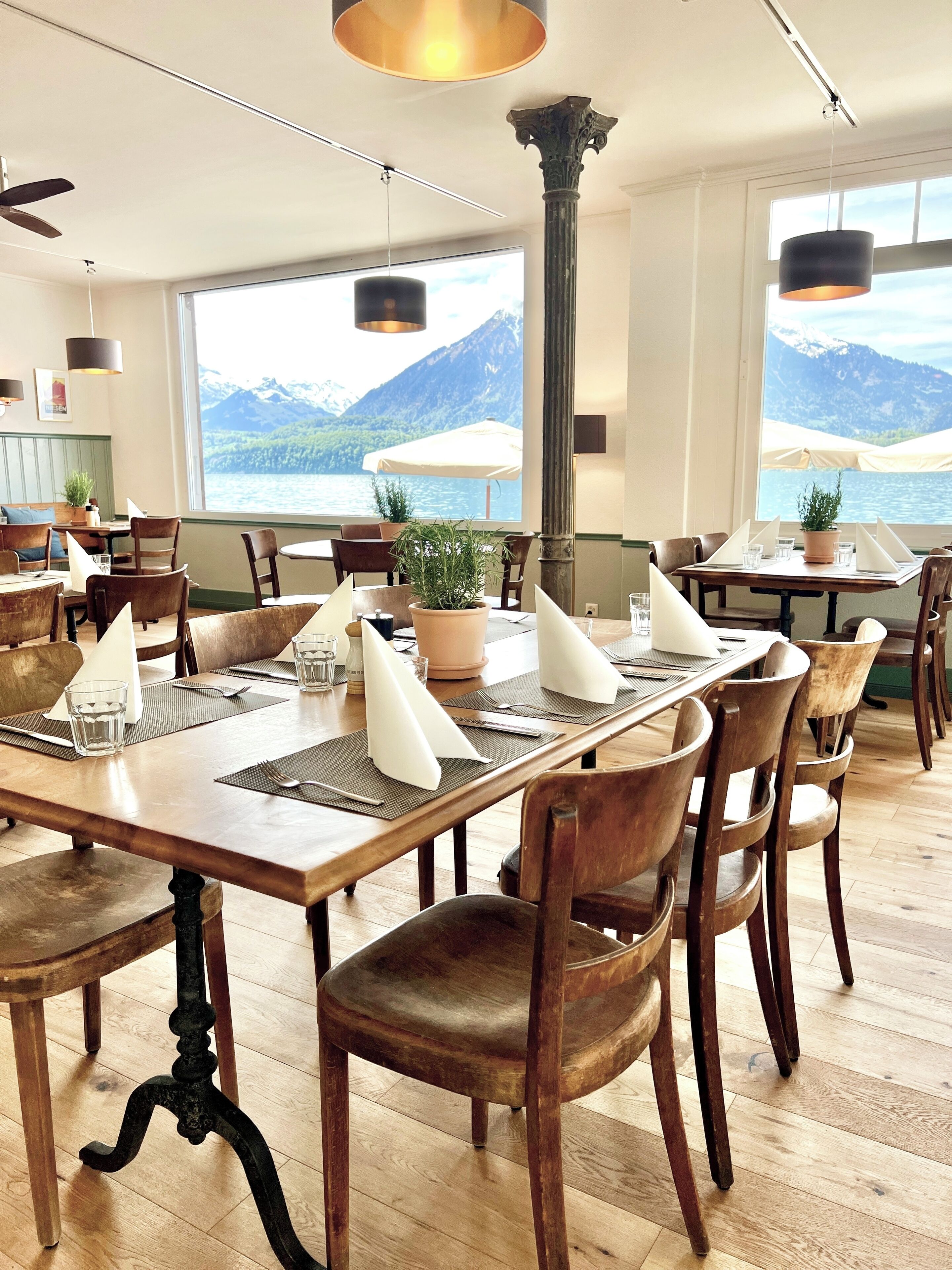 Un restaurant amb taules i caires de fusta, plantes en testos i vistes a les muntanyes i l'aigua.