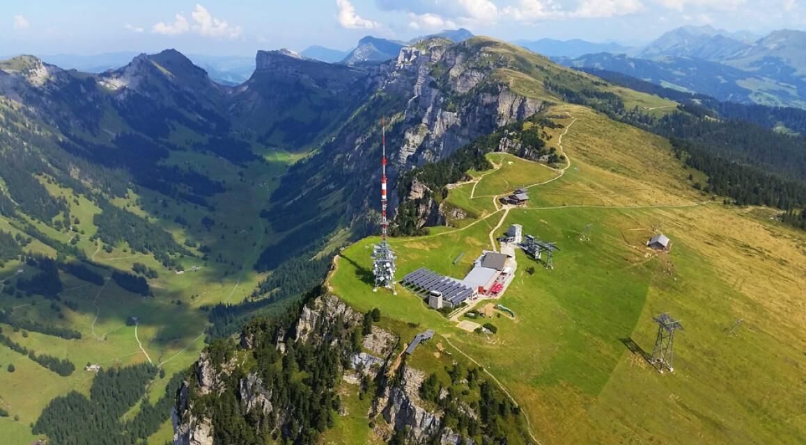 Eine Vogelperspektive auf eine gebirgige Gegend mit einem kleinen Gebäudekomplex, einschließlich eines hohen Turms, umgeben von grünen Feldern und bewaldeten Gebieten.