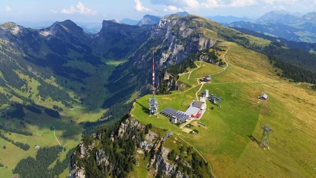 Vista aérea de una zona montañosa con un pequeño complejo de edificios, que incluye una torre alta, rodeado de campos verdes y zonas boscosas.