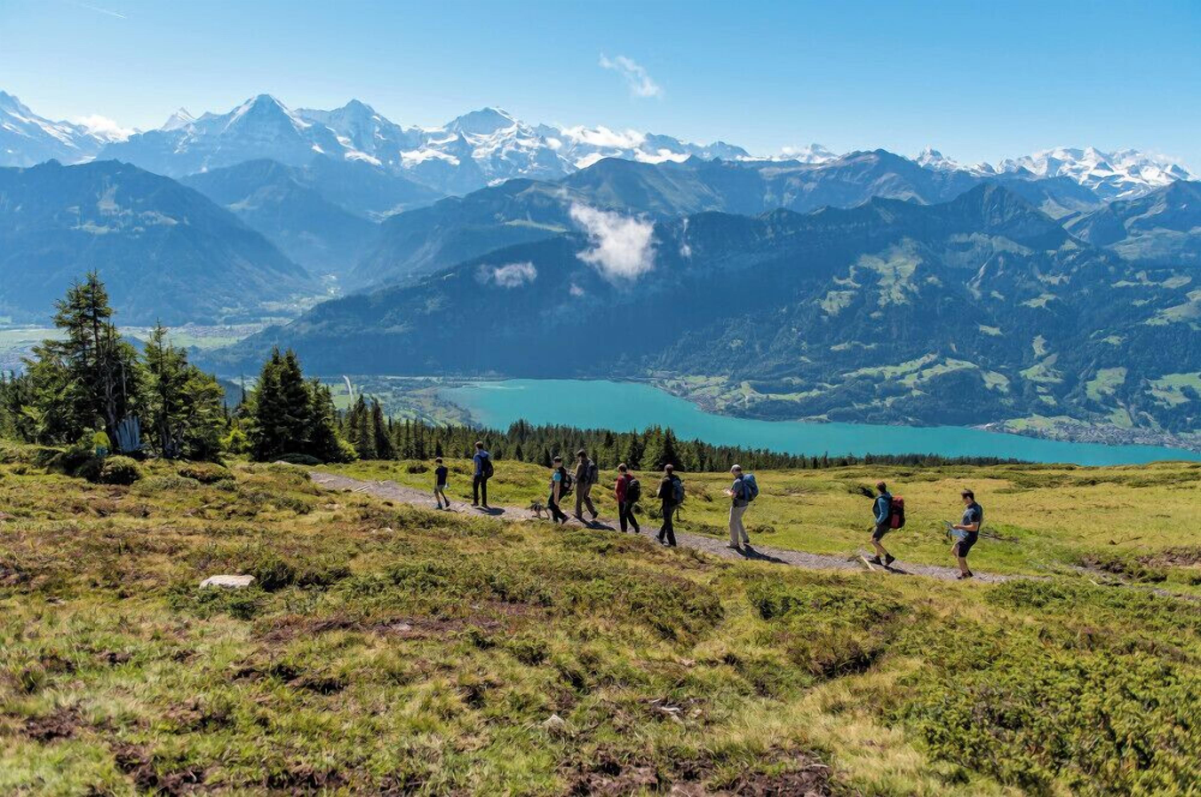 Um grupo de caminhantes numa trilha de montanha, com um lago e montanhas cobertas de neve ao fundo.