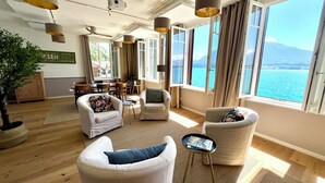 A spacious living area with multiple seating options, large windows offering a view of the sea, and a dining area with wooden furniture. - Boutique Hotel Niesenblick Self Check In (Oberhofen am Thunersee)
