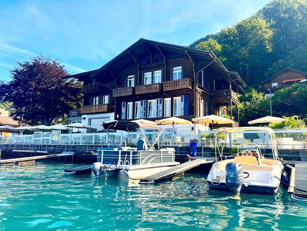 A wooden building with balconies, a dock with boats, and a clear sky. - Boutique Hotel Niesenblick Self Check In (Oberhofen am Thunersee)