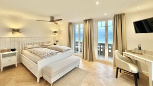 A bedroom with a bed, bedside tables, a desk, a chair, a TV, and a view of the sea. - Boutique Hotel Niesenblick Self Check In (Oberhofen am Thunersee)