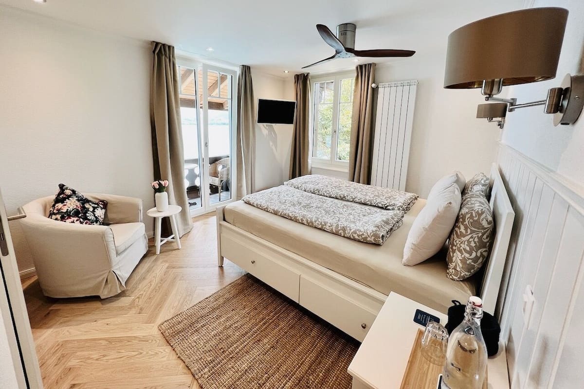 A modern bedroom with a bed, a chair, a small table, a ceiling fan, and a TV.