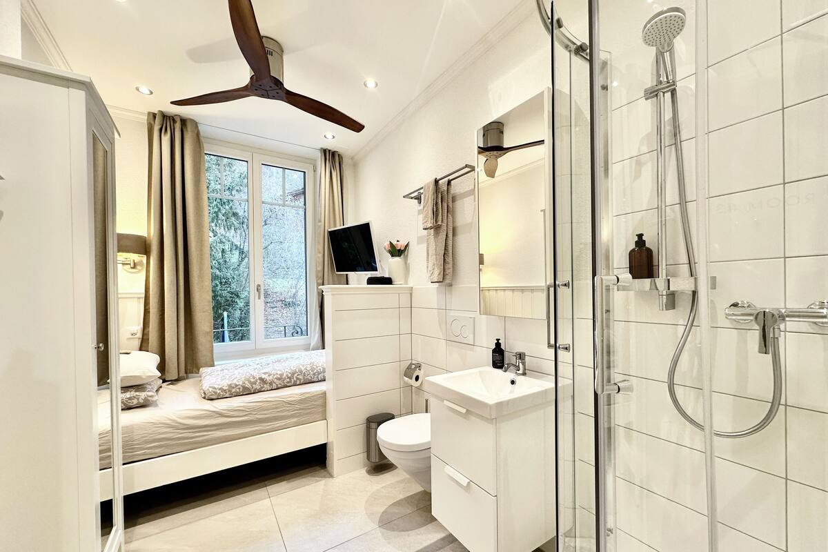 A modern bathroom with a shower, toilet, and sink. There is a window with curtains and a bed in the corner.