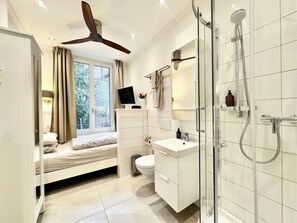 A modern bathroom with a shower, toilet, and sink. There is a window with curtains and a bed in the corner. - Boutique Hotel Niesenblick Self Check In (Oberhofen am Thunersee)