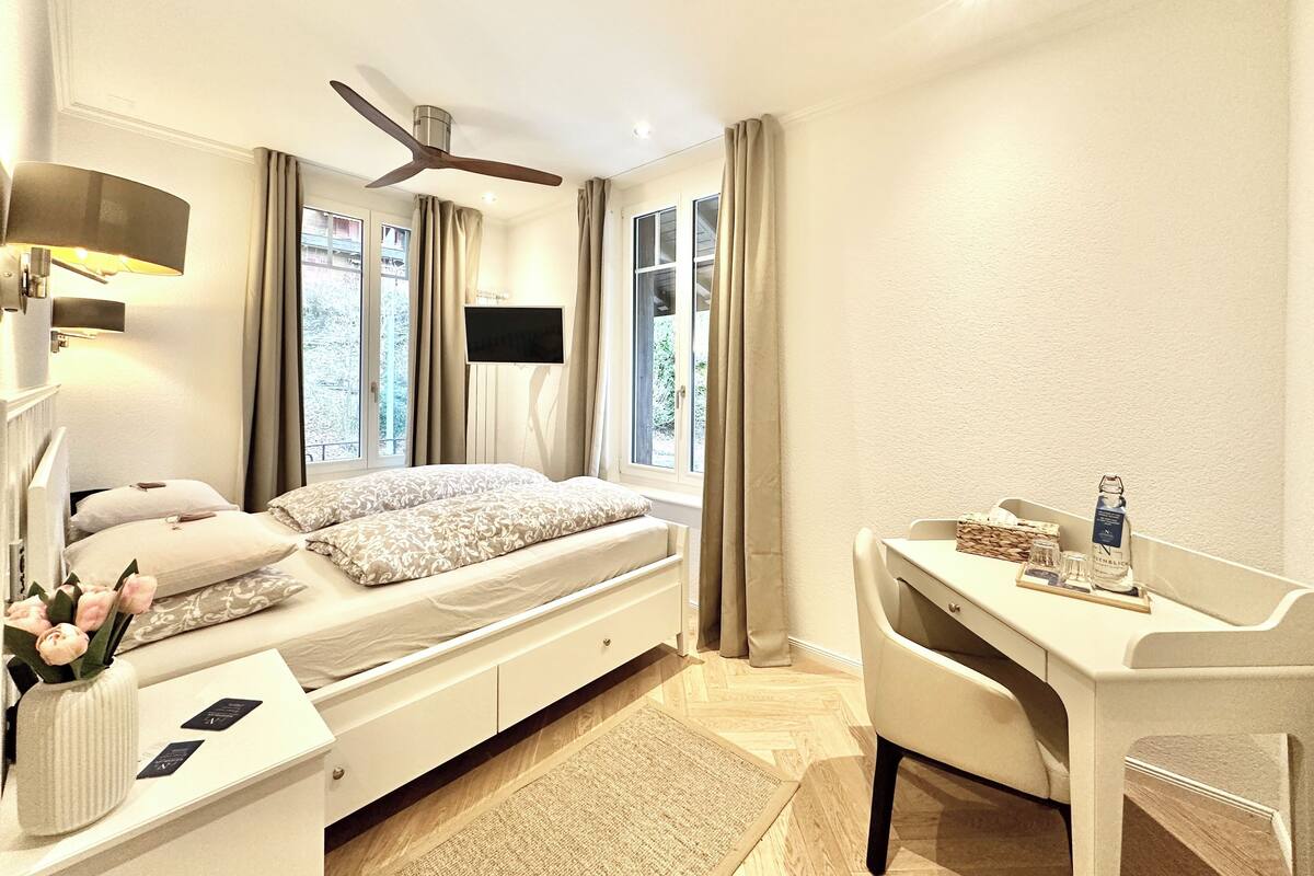 A bedroom with a bed, bedside table, desk, chair, TV, and a ceiling fan.