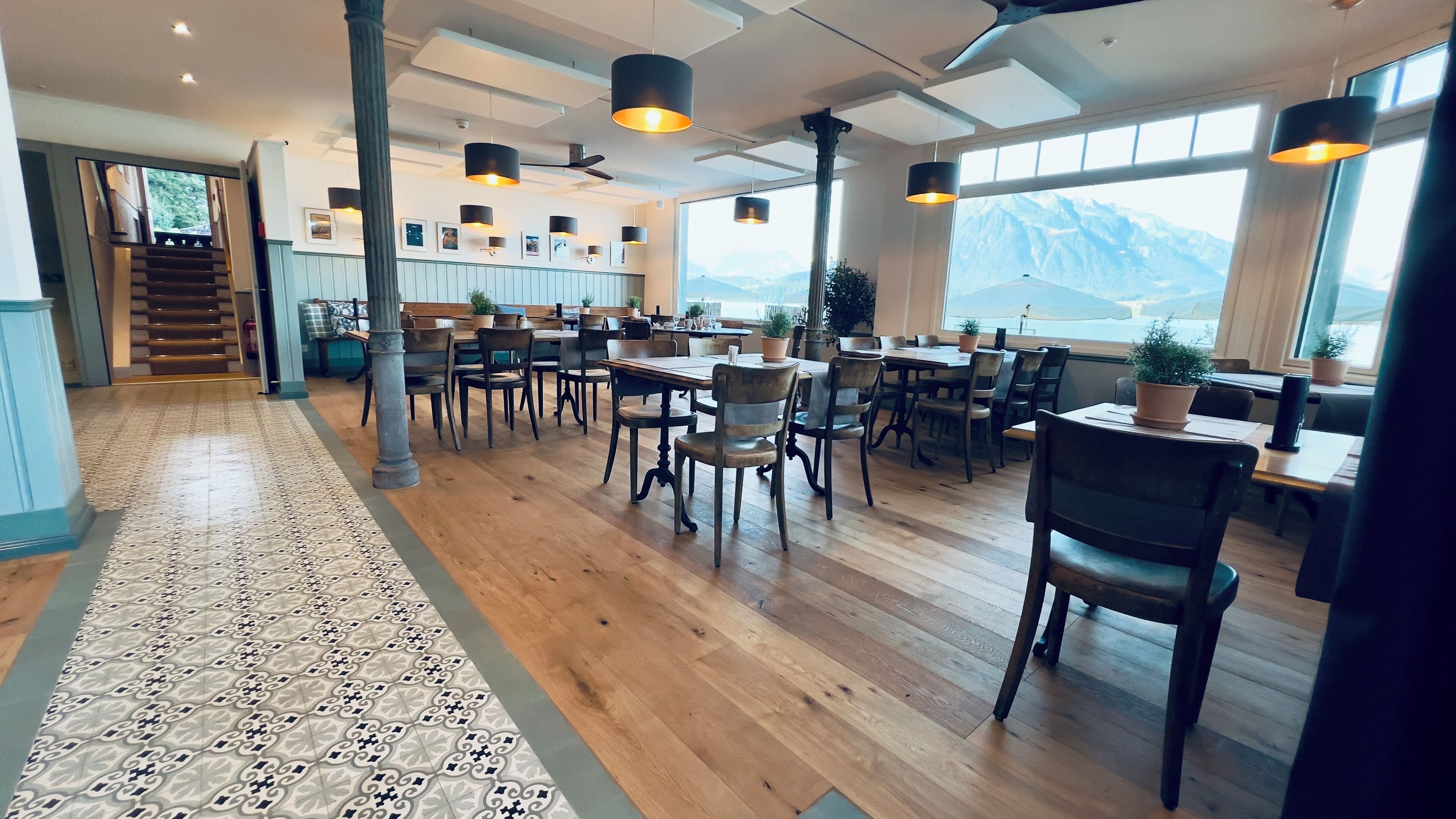 Un vaste espace dédié à la salle à manger, doté d’un plancher en bois, de nombreuses tables et chaises, d’un éclairage suspendu et de grandes fenêtres offrant une vue sur les montagnes.