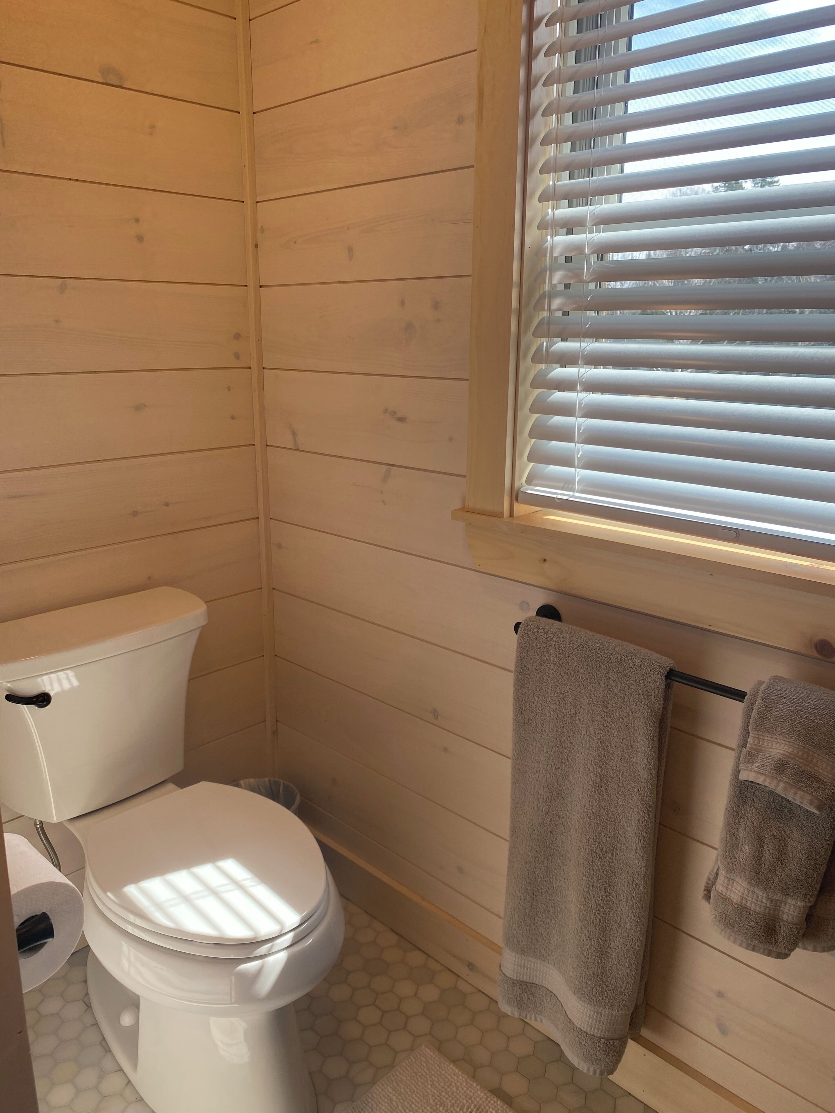 Un baño pequeño y funcional con paredes de madera, un inodoro, una ventana con persianas y dos toallas colgadas en un tendedero.