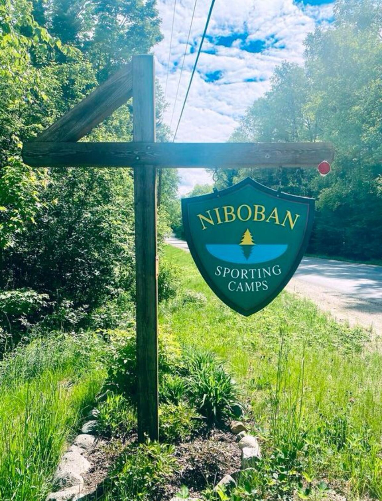 A sign for NIBOBAN SPORTING CAMPS with a logo, set against a backdrop of greenery and a road.