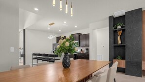 Un espace repas moderne avec une table et des chaises en bois, un vase rempli de fleurs et, au fond, une cuisine.