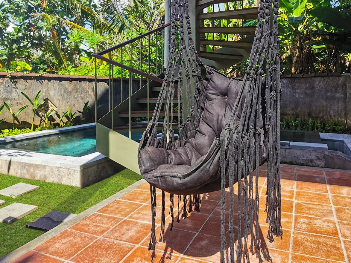 A hanging chair suspended from a wooden structure with a pool and garden in the background.