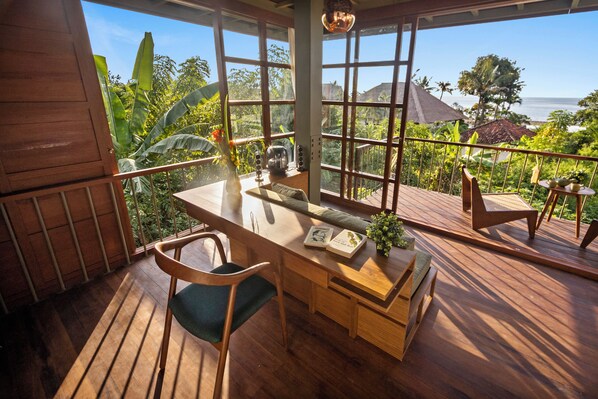 Una terraza de madera con una mesa de comedor y sillas, un pequeño rincón de estar, y grandes ventanales con vistas al océano y la vegetación tropical.