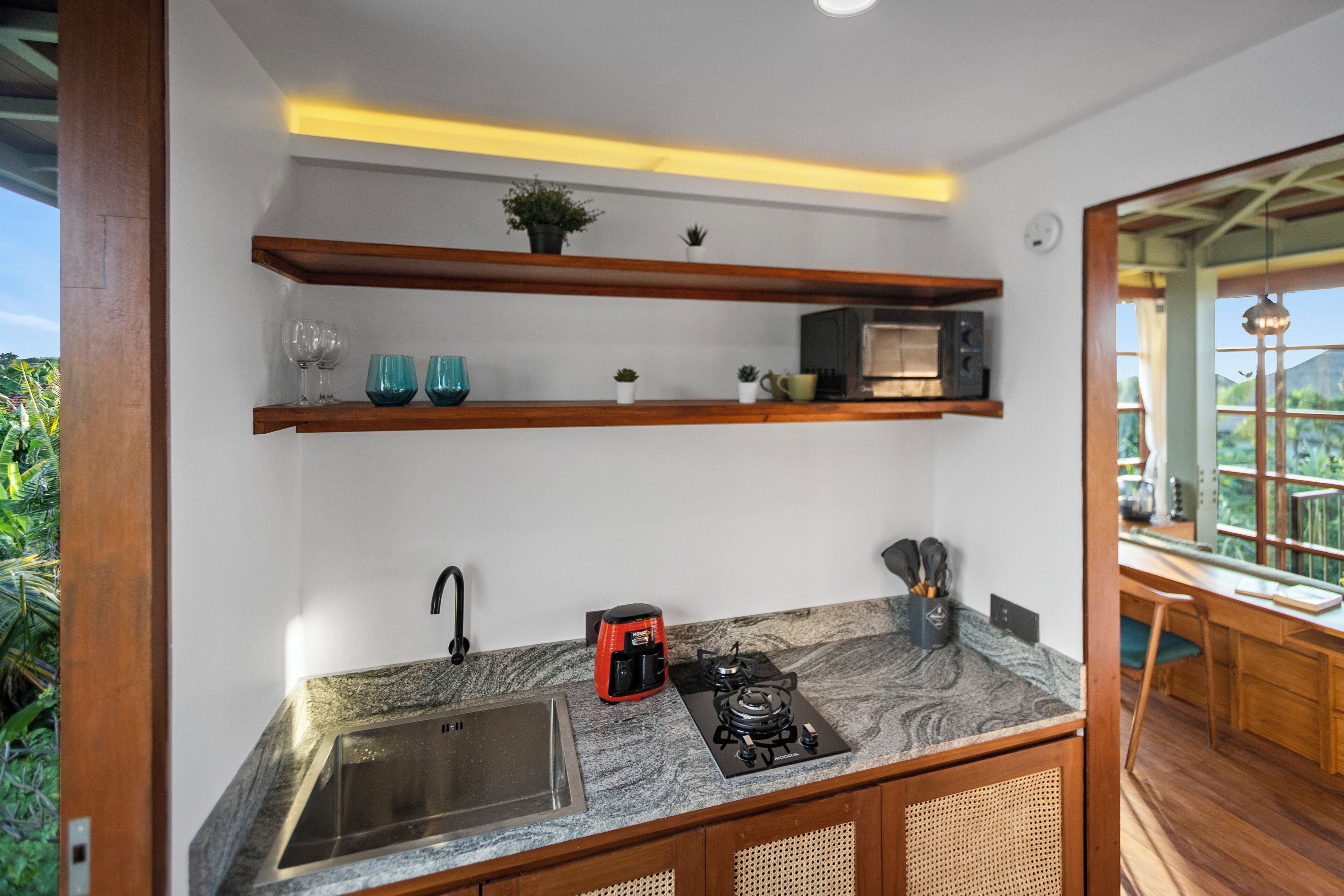 A kitchen with a granite countertop, stainless steel sink, and a built-in gas stove. There are wooden shelves above the countertop holding decorative items and a microwave.