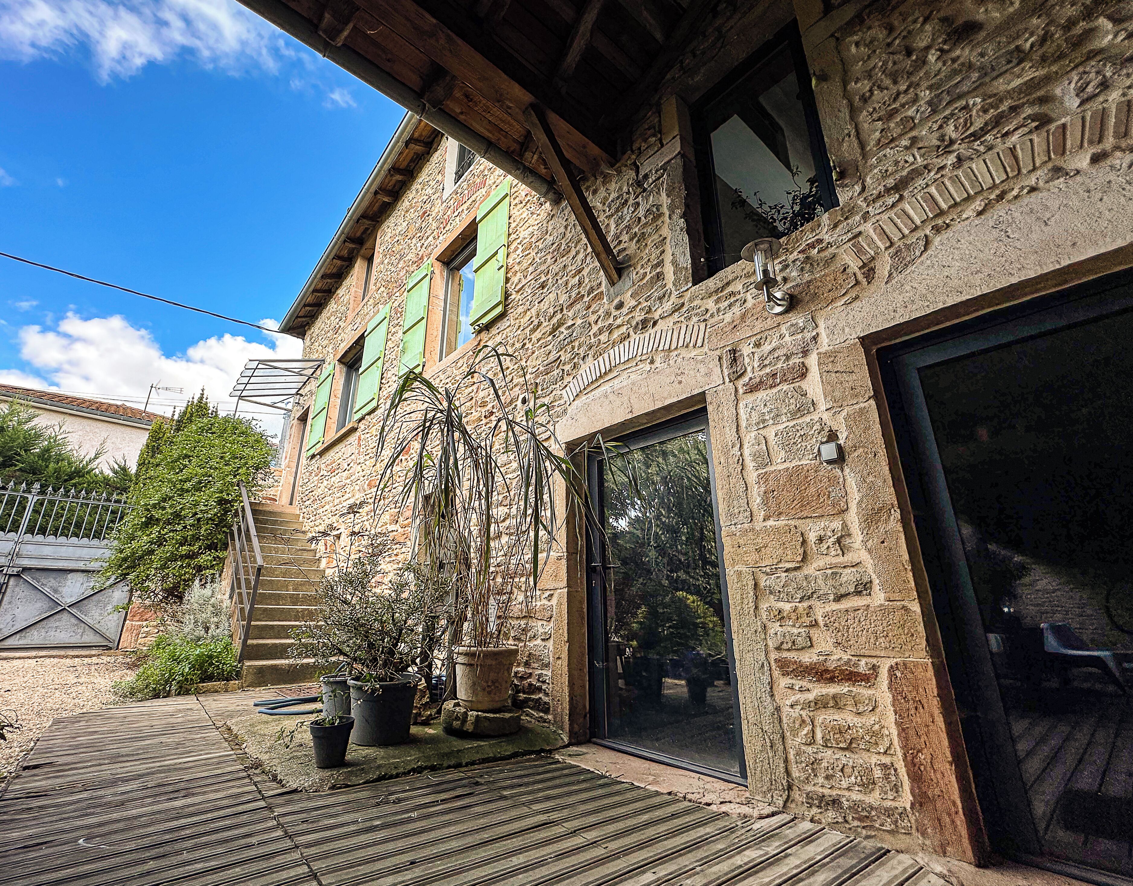 Ein Steinhaus mit einer Holzdielen-Terrasse, Pflanzgefäßen und einem großen Schiebeglastür.