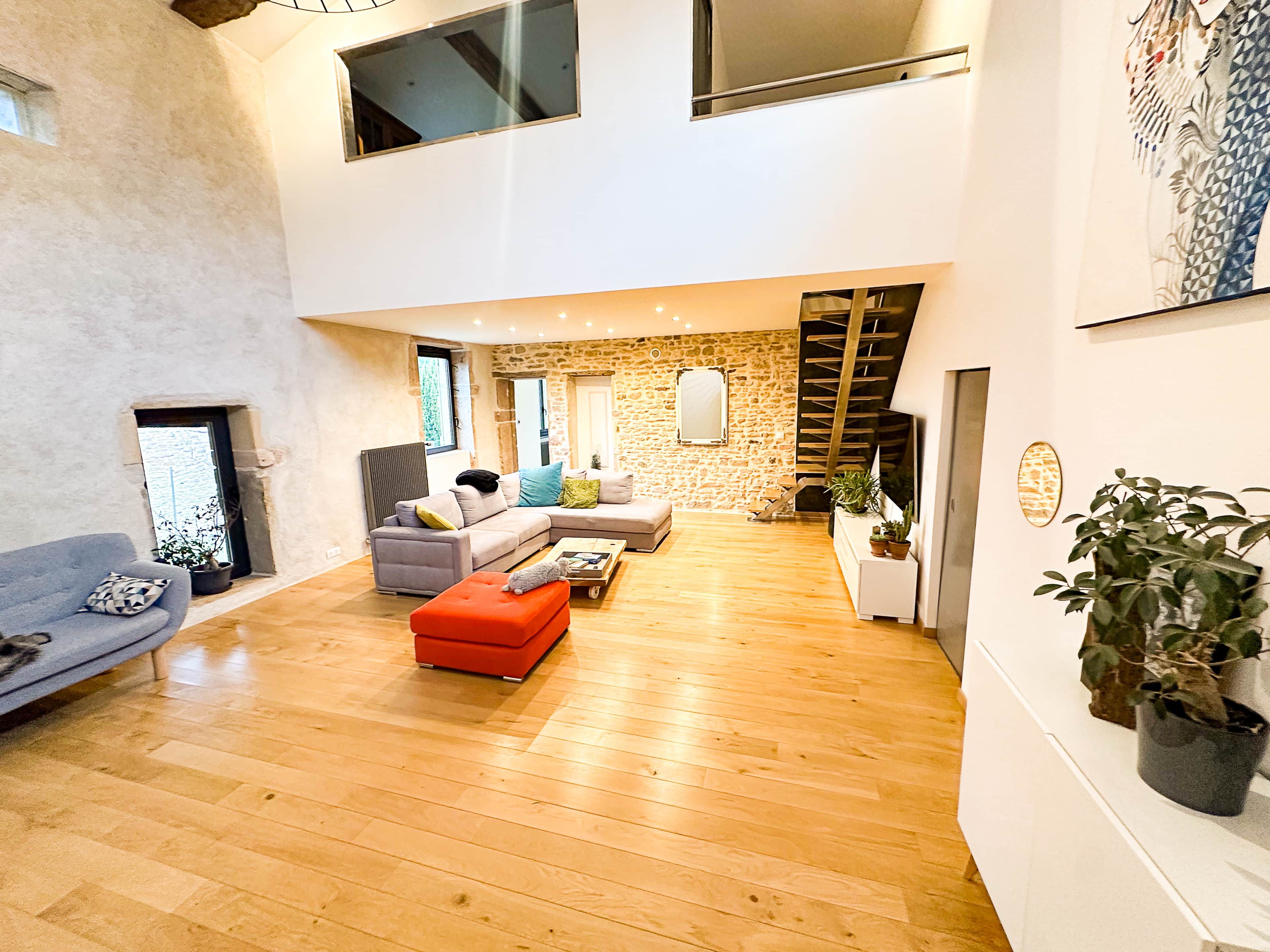 A spacious living area with a stone wall, a gray sofa, a red ottoman, and a wooden floor.