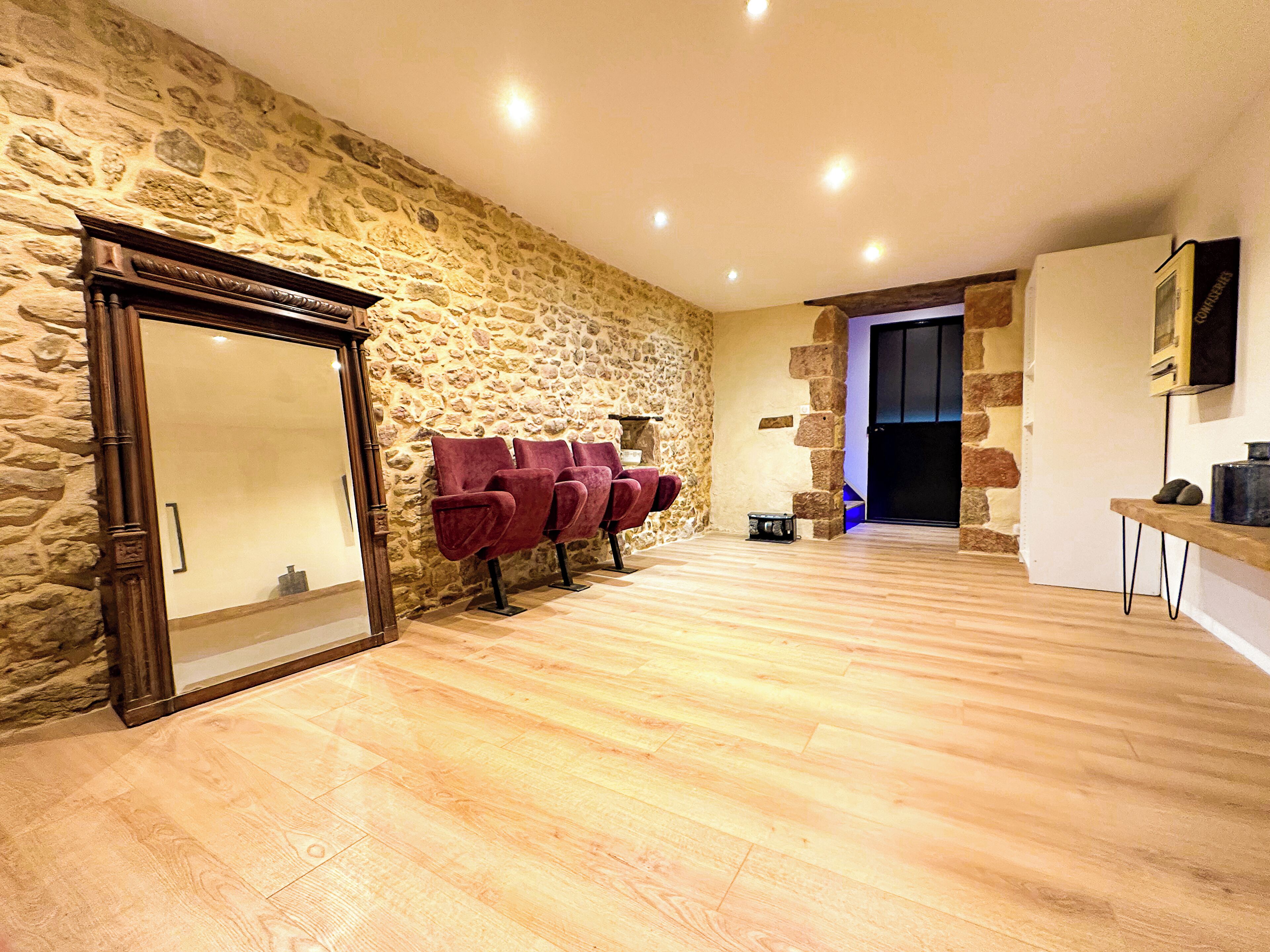 Una habitación con pared de piedra, suelo de madera y una puerta con espejo. Hay sillas rojas y una pequeña mesa con lámpara.