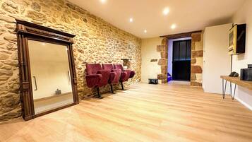 Una habitación con pared de piedra, suelo de madera y una puerta con espejo. Hay sillas rojas y una pequeña mesa con lámpara.