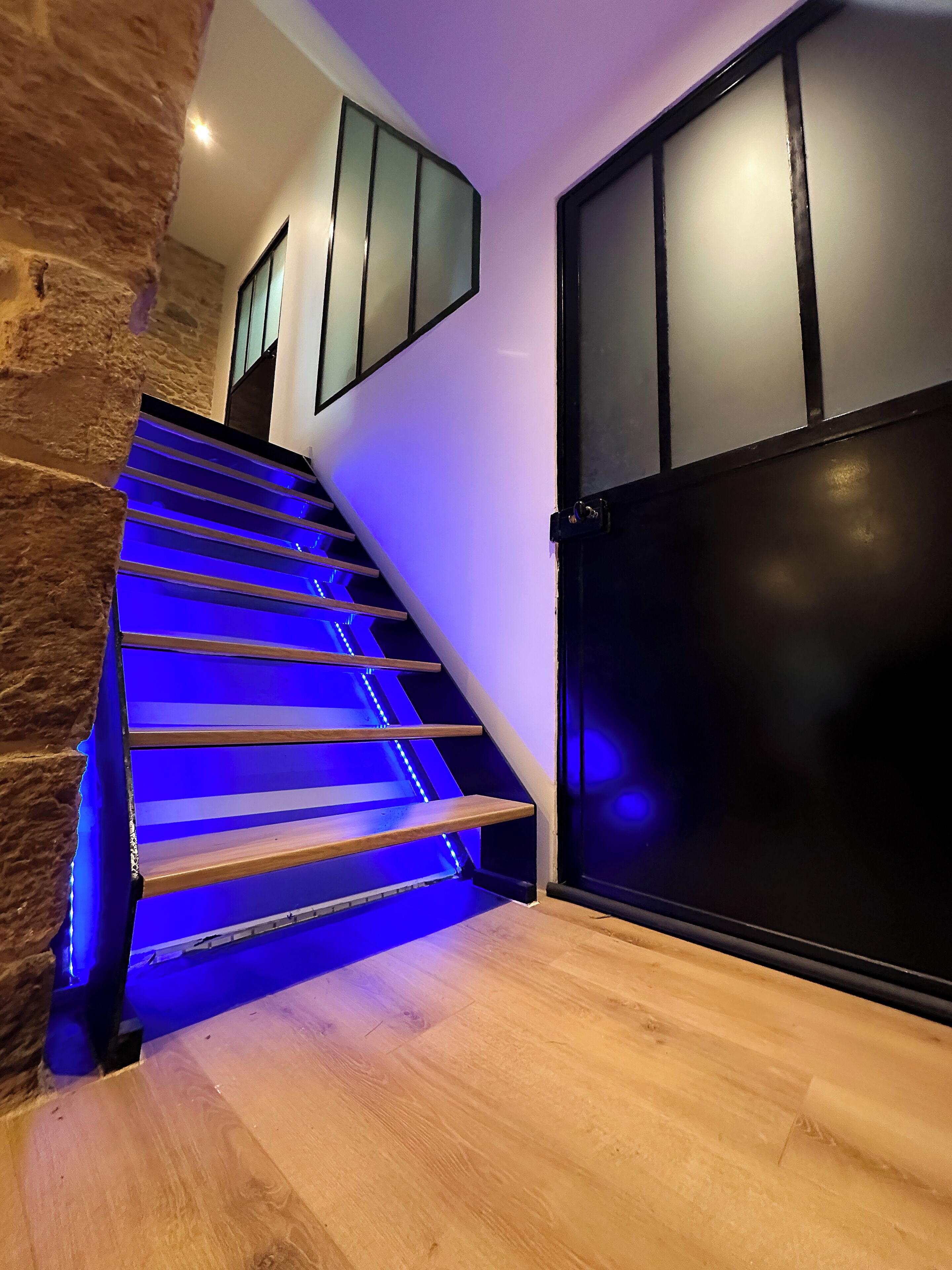 Een trap met blauwe LED-verlichting, een stenen muur en een zwarte deur.