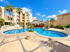 A swimming pool area with a thatched-roof structure, palm trees, and a multi-story building in the background.