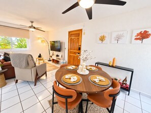 A dining area with a round table, four chairs, a ceiling fan, a TV, and framed artwork on the wall.