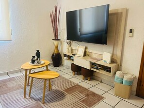 Un soggiorno con televisore a schermo piatto montato a parete, un piccolo tavolo rotondo con due sedie, un vaso decorativo e una libreria piena di libri e oggetti decorativi.