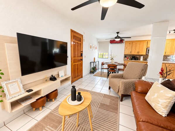 A living room with a flat-screen TV, a sofa, a coffee table, and a dining area with chairs.