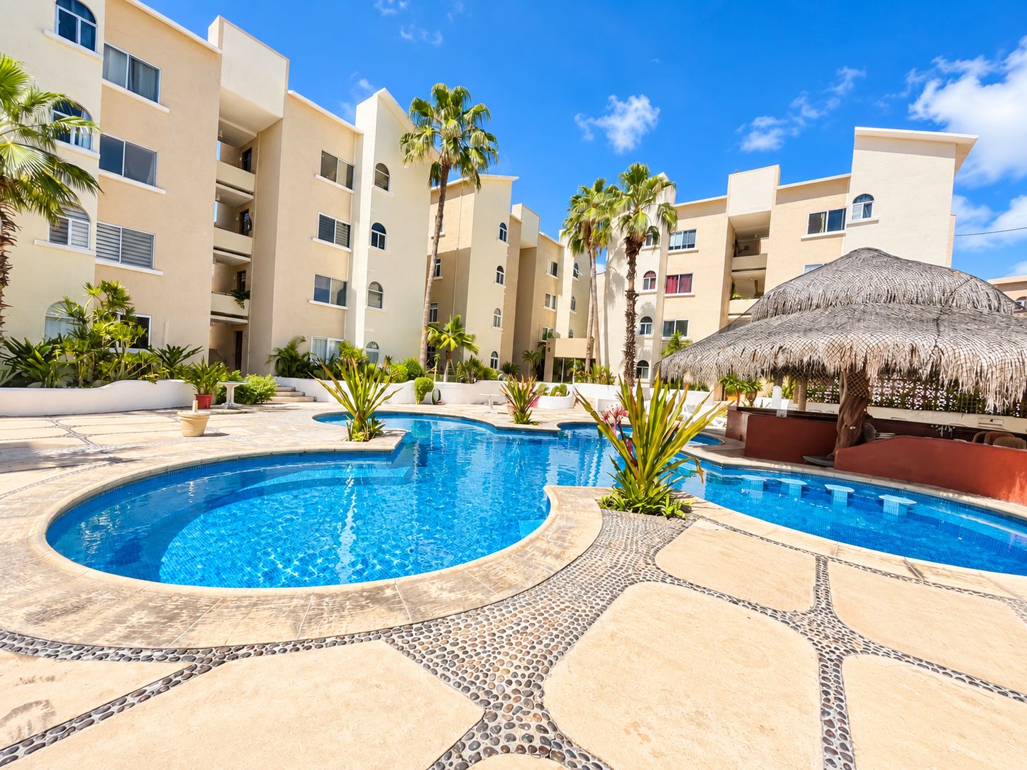A swimming pool with a pebble-lined edge, surrounded by palm trees and a thatched umbrella.