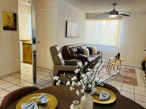 A living room with a brown leather sofa, a dining table with gold placemats, and a vase with white flowers.
