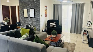Ein modernes Wohnzimmer mit grauem Sofa, einem schwarzen Sessel, einem Couchtisch und einem Essbereich mit Tisch und Stühlen.